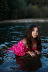 A woman with long dark hair sits in the water, wearing a pink dress, the water around her is blue