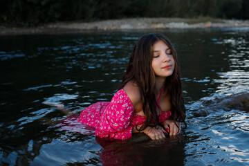 A woman with long dark hair sits in the water, wearing a pink dress, the water around her is blue