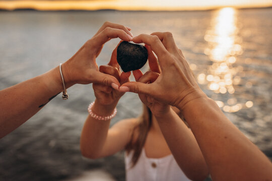 Two pairs of hands forming a heart shape around a stone at the beach during sunset, with a shimmering water background. Mother and daughter 
