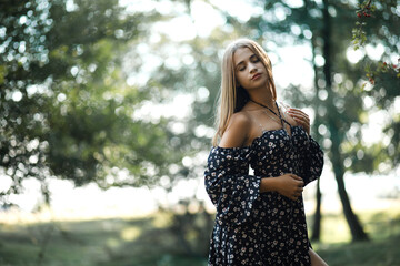 A woman in a dark dress with a floral pattern and a slit stands in a forest clearing on a summer...