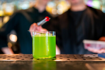 Bright green cocktail in a clear glass on bar counter, with blurred bartenders in background preparing garnish. Vibrant drink with frothy top in nightlife setting