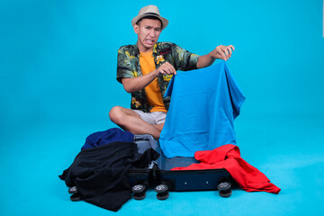Asian Man Looking Frustrated While Trying To Pack Clothes Into An Open Suitcase