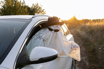 child in a white blanket and black glasses leaned out of a car window against an orange sunset....