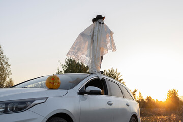 Unrecognizable child in white blanket, hat, black glasses stands on roof of white car at sunset....
