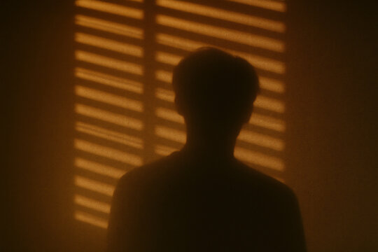 moody silhouette of man standing in front of window blinds with warm orange light and dramatic shadows