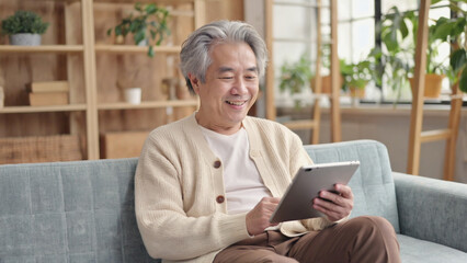 Happy senior Asian man with grey hair smiling and using a digital tablet while relaxing comfortably on a sofa at home.