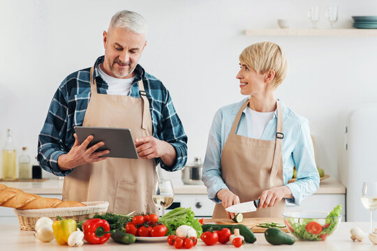 Homemade food and prepare holiday dinner for family at home together. Smiling senior man looks in tablet, wife prepares salad in modern kitchen interior with table with fresh vegetables, copy space - Powered by Adobe