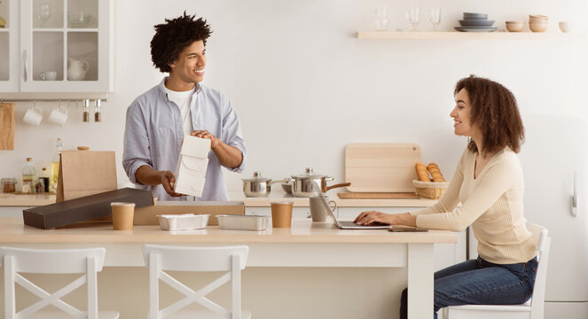 Modern work remotely, delicious takeaway food during covid-19 self-isolation. Happy young african american male takes out containers from box with order, lady works at laptop in kitchen interior