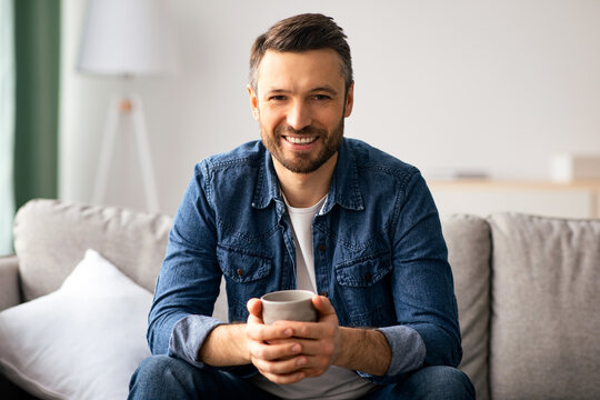 Relaxed handsome middle-aged man in casual outfit sitting on couch with mug of tea, caucasian bearded man drinking coffee in cozy living room at home, smiling at camera, copy space