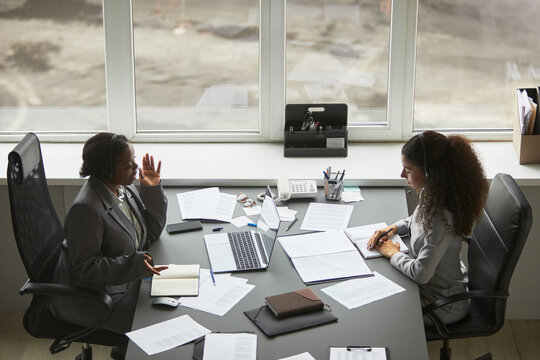 Two businesswomen, one Black and one biracial, sitting at office desk discussing documents and using laptop, both wearing headsets and engaging in professional conversation