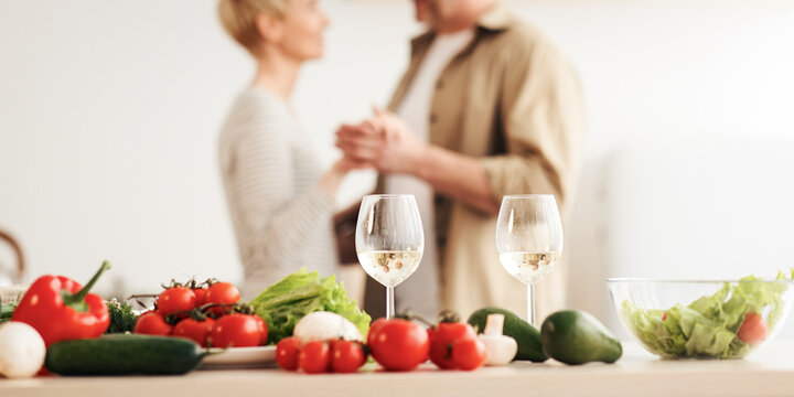 Romantic beautiful couple in modern cozy white kitchen room interior, celebrate holiday together at home. Glasses of wine on table with fresh vegetables, happy adult husband and wife dancing, blurred