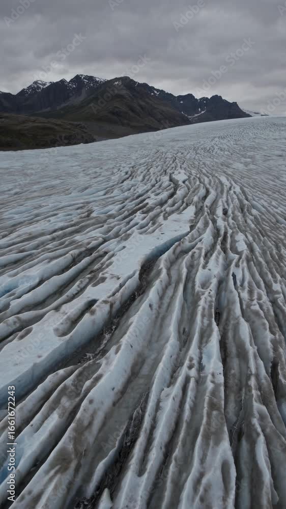 Poster aerial view of a vast glacier with rugged patterns, surrounded by mountains under a cloudy sky. perf