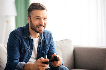Joyful middle-aged bearded man playing video game with joystick at home, sitting on couch in living room, having fun at weekend, copy space. Home entertainment, leisure, domestic lifestyle © Prostock-studio