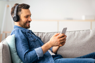 Positive bearded man with headset and mobile phone reclining on couch and smiling, home interior, side view, copy space, watching videos or listening to music, mobile application for smartphone