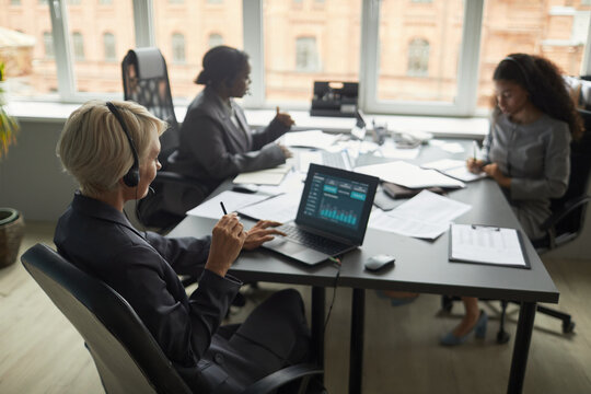 Caucasian middle aged woman wearing headset working at desk with laptop displaying financial charts, diverse colleagues collaborating in modern office setting