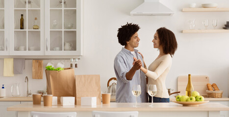 Family relationships, romantic dinner and food delivery. Happy millennial african american couple dancing in kitchen interior with eco food bags, paper dishes and glasses of wine on table, copy space