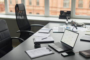 Modern office workspace featuring open laptop, smartphone, documents, pen and coffee cup on desk near large window, empty ergonomic chairs and additional laptops in background
