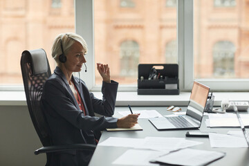 Caucasian middle aged woman wearing headset sitting at desk working on laptop, taking notes during business call in modern office environment with large windows in background