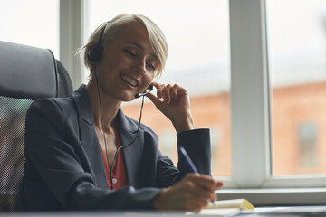 Caucasian middle aged woman smiling while wearing headset, sitting at desk, holding pen and writing...