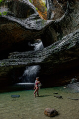 A young Caucasian woman stands in a shallow pool of water beneath a rocky overhang with a waterfall cascading in the background. The scene captures natural beauty in the Caucasus region.