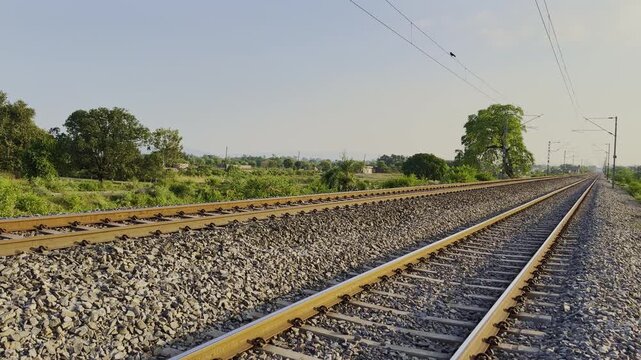 A serene railway scene unfolds with parallel tracks surrounded by gravel and lush greenery, set against a natural backdrop of trees and bushes, with power lines above