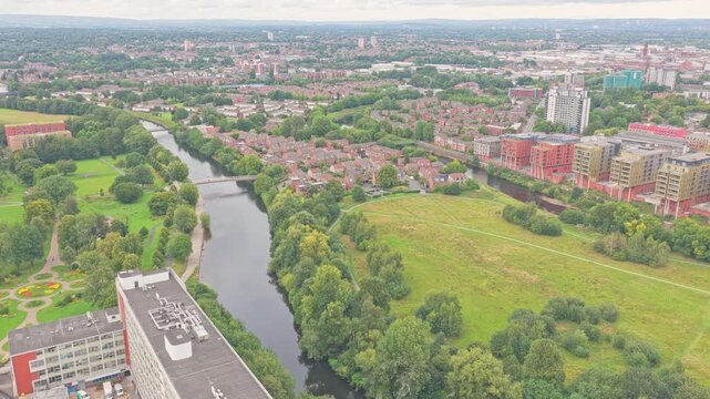 Aerial static shows Salford Meadows and River Irwell, featuring the curved silhouette of Calatrava&rsquo;s Trinity Bridge connecting Salford to Manchester amid green parkland and urban backdrop.
