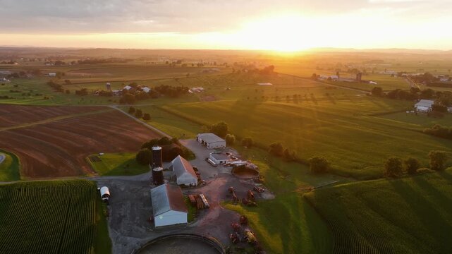 Large American farmstead with silo storage, barn and fields during bright golden sunset. Circular pond in rural suburb area of town. Aerial wide shot. Midwest, USA. Peaceful countryside scene.