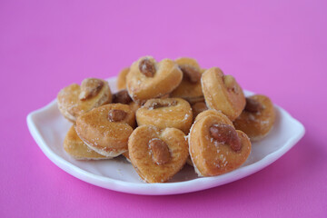 Heart-shaped cashew nut cookies, also known as Singapore cookies, arranged on a white plate over pink background. Golden, crunchy, and sweet, perfect for bakery, dessert, or snack concept.            