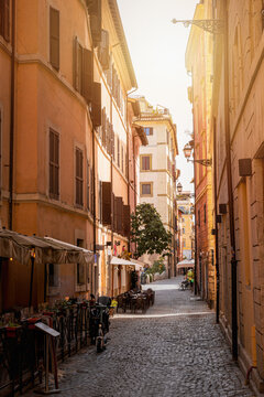 Narrow cobblestone street in Trastevere, Rome, Italy, with cafes, tables, warm sunset light and colorful buildings