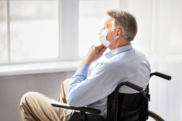 Disabled seniors during covid quarantine. Handicapped elderly man in face mask looking out window at home, feeling lonely and depressed, staying indoors at times of coronavirus lockdown