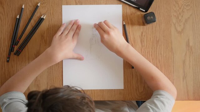 The creative process of a child drawing a character from imagination. Kid's hands erase and redraw a pencil sketch. Homeschooling, art class and skill development, top down view.