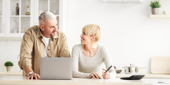 Smiling senior couple spouses use laptop, calculator, new technology paying banking bills online. Happy modern male and lady manage domestic finances expenditures considered family budget in kitchen