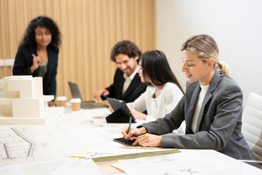 Businesswoman focused on working with a digital tablet while her colleagues having a meeting in the background, discussing on house model in modern conference room
