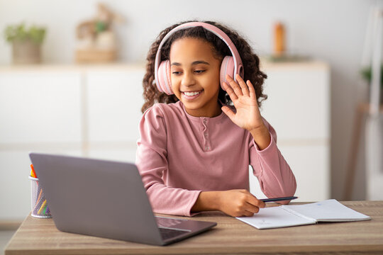 Home education. African american girl having video conference with teacher or friends from home, using laptop and waving at computer screen. Happy schooler wearing wireless headphones and smiling