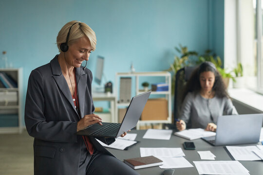 Caucasian middle aged woman wearing headset using laptop while standing in modern office, young biracial woman working at desk with documents and computer in background - Powered by Adobe