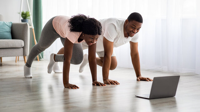 Sporty young african american couple in sportswear exercising together in living room, having online fitness class, video call with trainer from home while covid-19 pandemic, using laptop, copy space