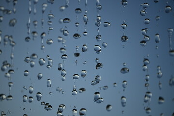 Macro photo of water droplets falling from a curtain fountain, captured in mid-air against a clear sky. Close-up showing fluid texture, shape, motion, and transparency.