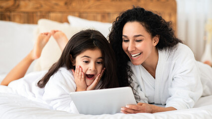 Emotional young mom and little girl laying on bed with digital tablet, playing games together and winning, wearing white bathrobes. Happy mother and daughter having beauty day together at home