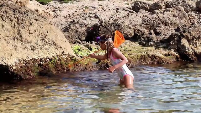 La Pineda, Spain - June 09, 2023: Young girl in white swimsuit exploring rocky shoreline with a net, discovering marine life in clear water