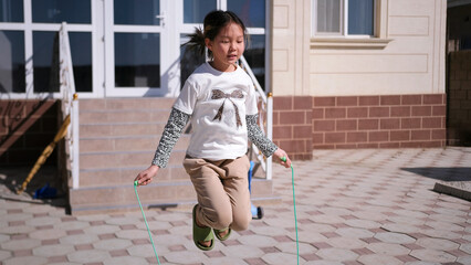 A teen girl jumps rope in front of her house. Physical activity of a teenager. Girly games. The child is engaged in a fun activity in the fresh air © chekart