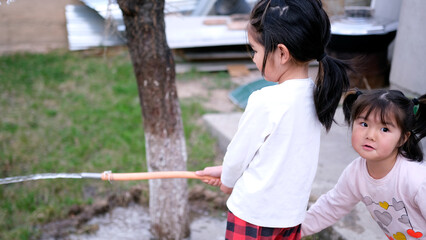 Two funny sisters are watering plants in the garden with a hose. Two little Asian girls are playing...