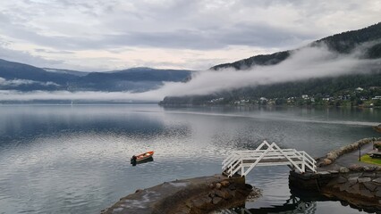 Tranquil misty morning over Innvik fjord in Norway, with a small red boat floating near a white wooden pier. Scenic travel and tourism destination, perfect for relaxation and nature lovers