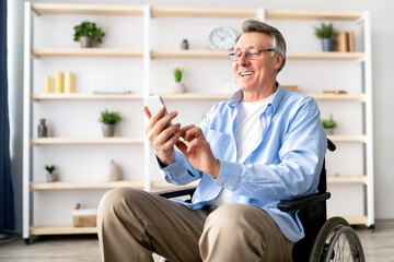 Happy disabled older man in wheelchair using cellphone, browsing web or watching movie at...