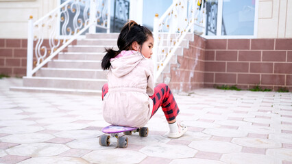 A little girl is sitting on a skateboard in front of the house. An Asian child learns to...