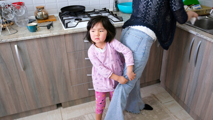 A little daughter does not take a single step away from her mother. A woman is washing dishes in...