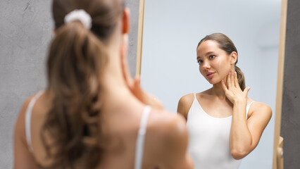Woman with long brown hair in white tank top examines her healthy skin in a mirror, showcasing a serene atmosphere and wellness concept, reflecting self-care and beauty routines