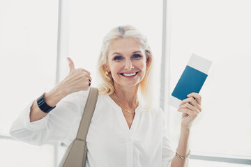 Smiling woman holding travel ticket in bright airport terminal showing thumbs-up expressing...