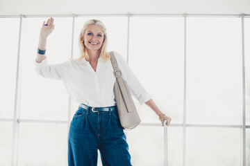 Elegant mature woman waving and smiling, holding a suitcase handle, waiting in an airport terminal...