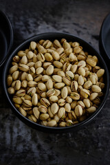 A macro shot of roasted salted pistachios in a black ceramic bowl, captured in a dramatic, moody style on a dark, textured surface.