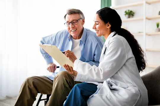 Young doctor asking senior impaired male patient in wheelchair to sign insurance policy at home. Handicapped elderly man putting his signature under surgery consent form, reading medical document